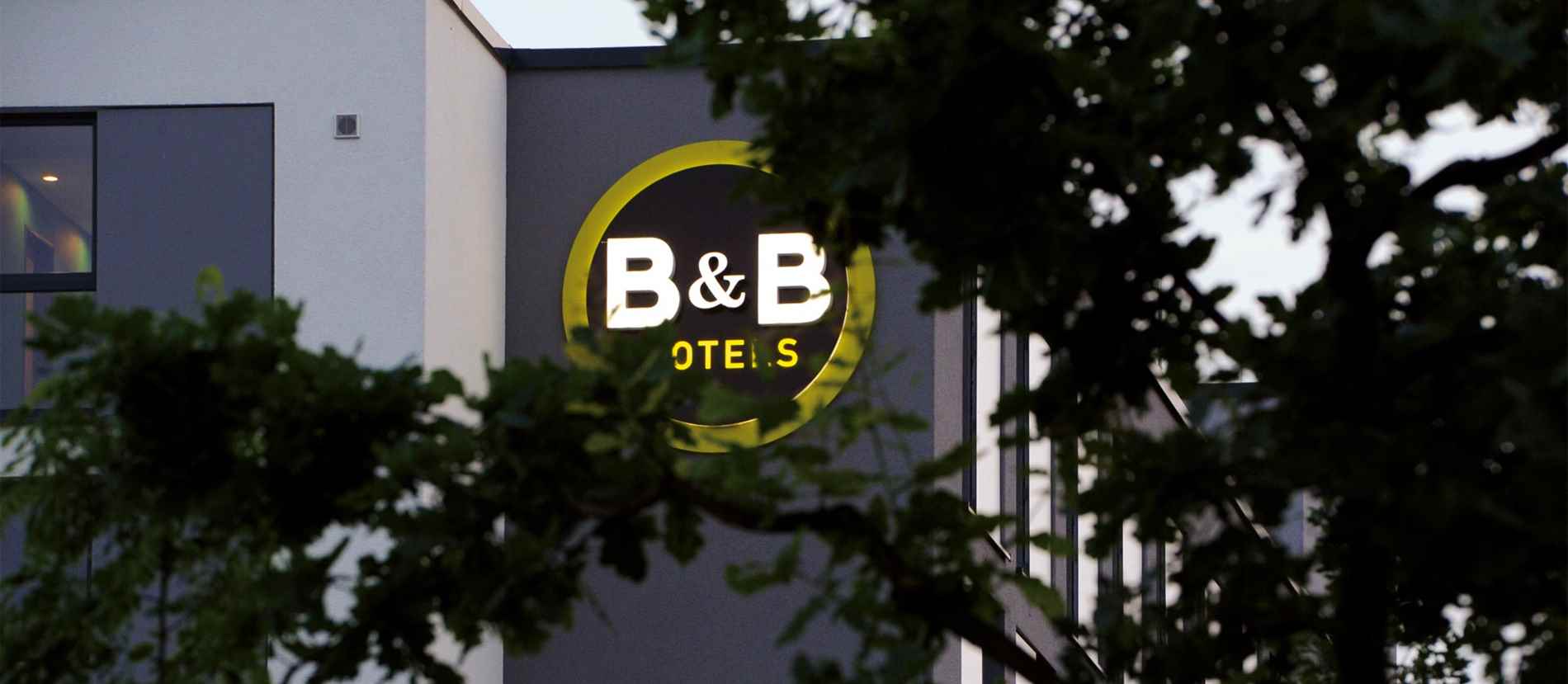 Der Blick geht durch die Äste eines Baums in der Dämmerung. Dahinter ist eine große Lichtwerbeanlage zu sehen in gelb und weiss. Die Werbeanlage weist darauf hin, dass es sich um ein B&B-Hotel handelt. Die Leuchtschrift ist von guttenberger+partner Lichtwerbung.