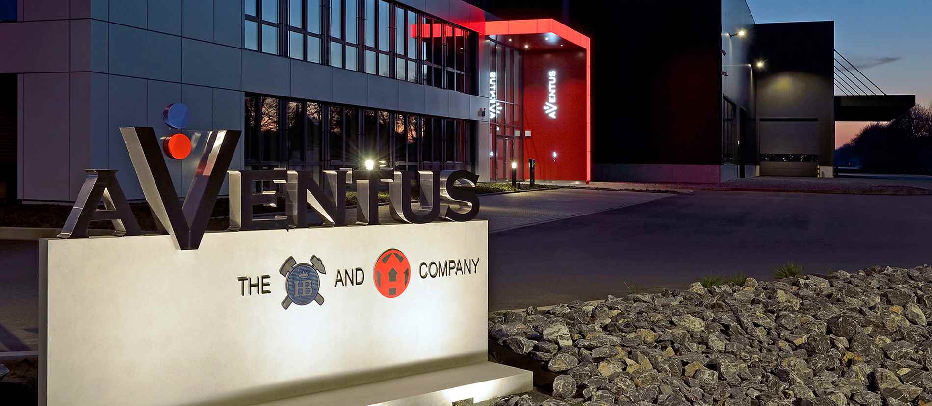 Das Bild zeigt eine repräsentative Lichtwerbeanlage an einem modernen Gebäude in Warendorf, gestaltet von Guttenberger & Partner. Im Vordergrund befindet sich ein beleuchtetes Schild mit dem Schriftzug "AVENTUS" und Logos darunter. Die Fassade des Gebäudes ist in modernen Materialien gehalten, mit großen Glasflächen und einer rot beleuchteten Ecke, die das Logo "AVENTUS" trägt. Das Gebäude und die Lichtwerbeanlage sind stilvoll und professionell gestaltet, was den hochwertigen Anspruch von Guttenberger & Partner unterstreicht.