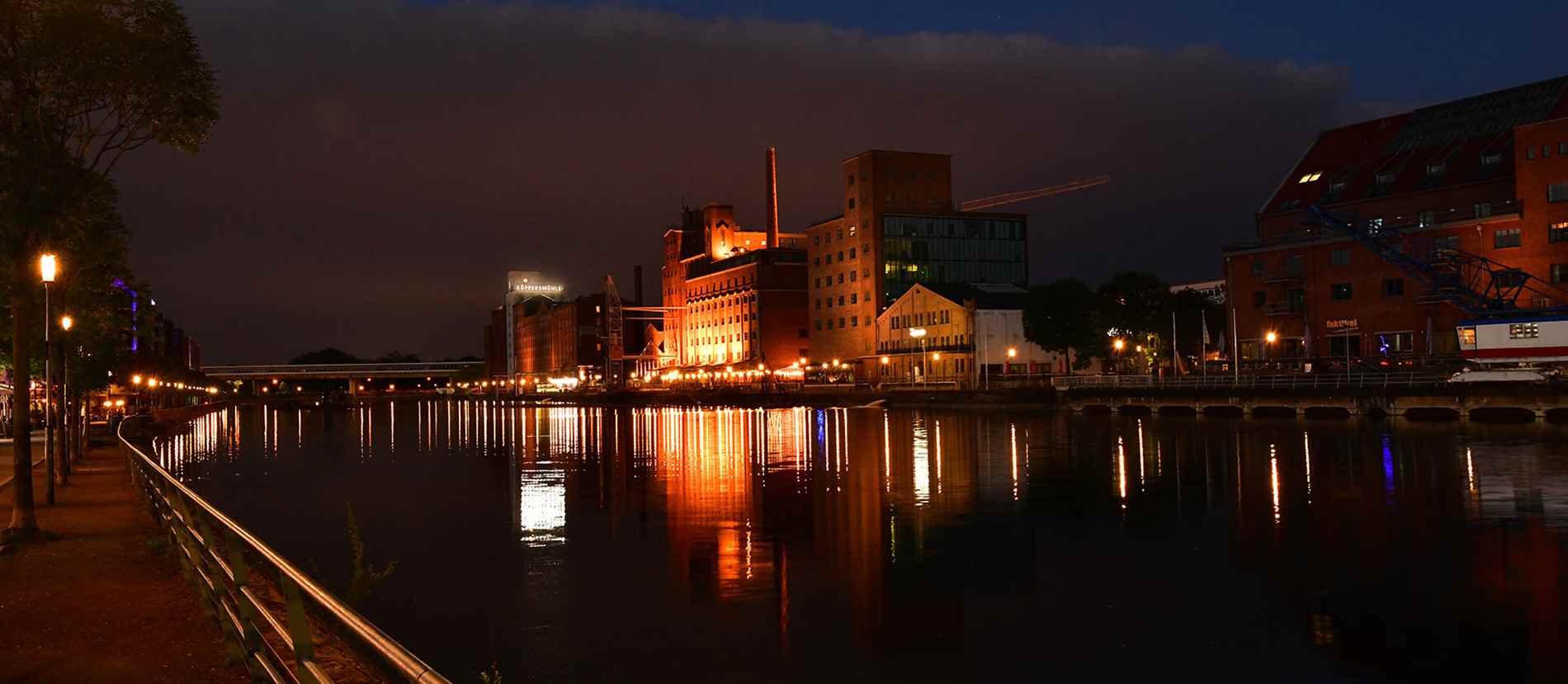 Beleuchtete Uferpromenade in Duisburg bei Nacht mit Blick auf das KÜPPERSMÜHLE-Museum. Die reflektierenden Lichter der historischen Gebäude und die stimmungsvolle Beleuchtung der Umgebung schaffen eine malerische Atmosphäre entlang des Wassers.