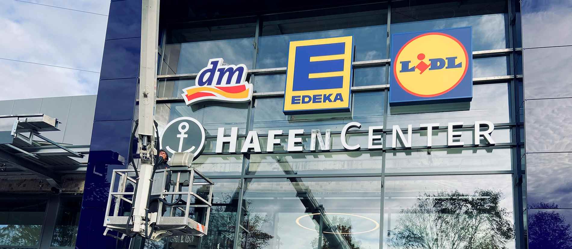 Lichtwerbung an der Glasfassade des HafenCenters in Mülheim Speldorf. Die modernen Leuchtlogos von dm, EDEKA und Lidl heben sich hervor und ziehen Kunden an. Ein Techniker arbeitet an der Installation der hochwertigen Lichtwerbung, die das Einkaufszentrum in Szene setzt.