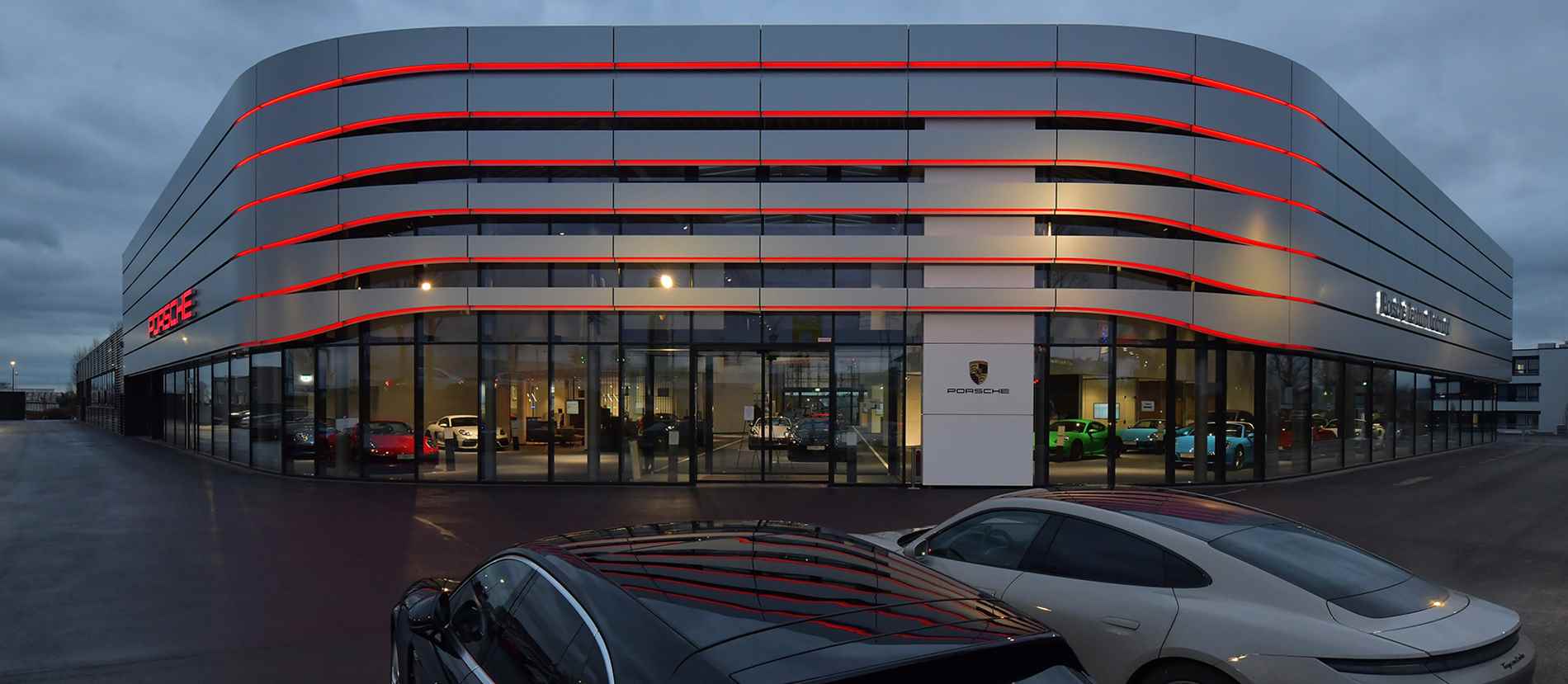 Die elegant geschwungene Fassade des Porsche-Zentrums in der Dämmerung, akzentuiert durch charakteristische rote LED-Leuchtstreifen, die die Konturen dynamisch hervorheben. Die transparente Glasfront bietet einen verlockenden Blick auf die Palette ikonischer Porsche-Modelle, während die reflektierenden Oberflächen der geparkten Fahrzeuge im Vordergrund das avantgardistische Design und den exklusiven Charakter der Marke unterstreichen.