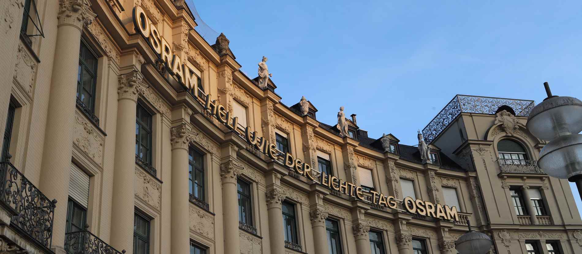 Historische Fassade eines Gebäudes am Stachus in München, verziert mit Säulen und kunstvollen Details. Der goldene Schriftzug ‚OSRAM HELL WIE DER LICHTE TAG‘ ziert die Oberseite des Gebäudes. Auf dem Dach sind mehrere Statuen zu sehen. Das Bild wird von modernen Straßenlaternen im Vordergrund ergänzt und fängt die Kombination aus traditioneller Architektur und moderner Werbung ein.