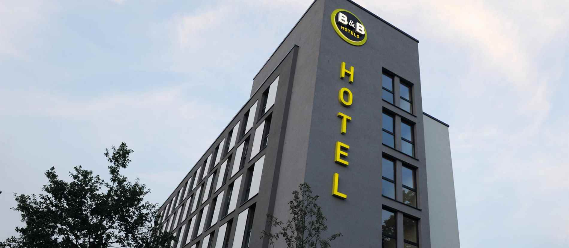 Gelbes Leuchtschild mit dem Wort "HOTEL" an der grauen Fassade eines mehrstöckigen Gebäudes. Darüber rundes Logo mit "B&B HOTELS". Gestaltet von Guttenberger und Partner, deutscher Anbieter für Leuchtreklame.